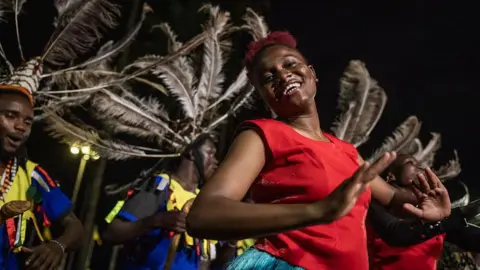 Getty Images Kenyan dancers wearing bight costumes adorned with feathers smile broadly as they perform under a dark night sky.