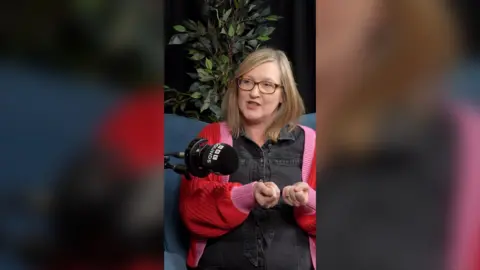 A woman with a plant behind her sits on a chair. She's wearing tortoise-shell coloured glasses and a red and pink cardigan.
