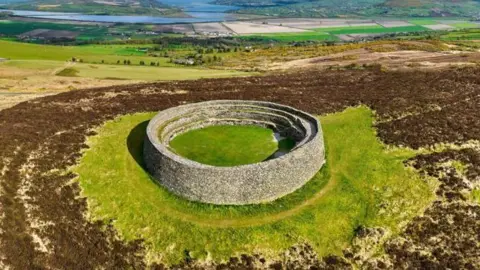 A ring fort made of stone sits on a grassy mound. it is surrounded by grown gorseland. In the background a blue body of water can be seen. 