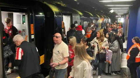 Sunderland AFC fans, many wearing red and white-striped kit, line up to board a train on a busy station platform in Sunderland.