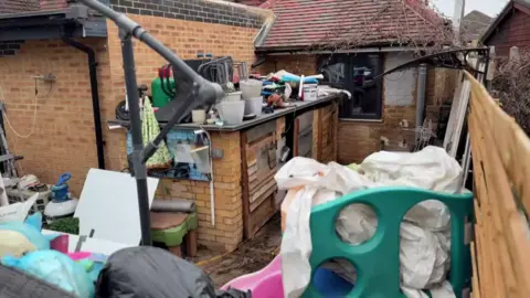A messy outdoor scene in the backyard of Save A Paw animal rescue centre. There are lots of buckets, bags and sheets all piled up behind a brick property.