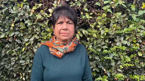 Tara Mistry standing in front of a green bush. She has short dark-brown hair styled in a pixie cut and is wearing a green jumper with an orange and green paisley scarf around her neck.