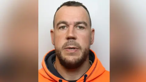 Daniel looks at the camera with a blank expression. He wears a bright orange hoodie and has a dark beard and thinning short dark hair.