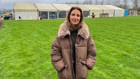 A woman with dark blonde and a brown coat standing on a football ground. 
