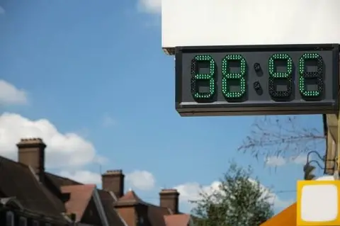 Getty Images A digital thermometer in London showing the temperatures during the UK heat wave. It reads 38 °C. In the background there are trees and houses, but only the roofs and chimneys are visible. 