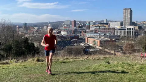 Mark Ancel / BBC Dot Caisterton Sheffield se ejecuta en arcilla de madera, donde entrenó en el fondo del centro de la ciudad detrás de él. Lleva una camiseta Red Parkroon T, pantalones cortos azules y un entrenador rojo. Es un sol, día de primavera.