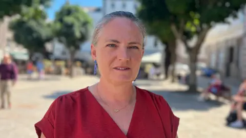 Deputy Hilary Jeune is standing in Jersey's Royal square, she's wearing a red top with a V-neck and blue earrings.