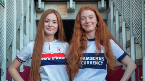 Tom Banks and Paul Thompson Grace and Eve are standing in an entrance tunnel at a stadium and wearing different white and blue Middlesbrough shirts. They have long ginger hair and they are both smiling at the camera. 