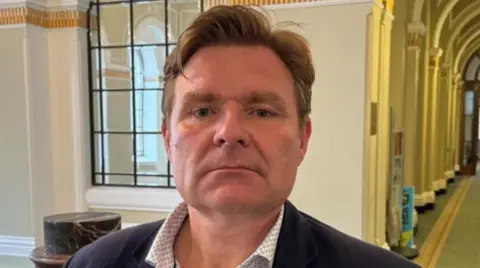 A man wearing a shirt and dark jacket is standing in a corridor. He has brown hair with a side parting.