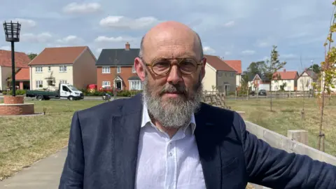 Ben Schofield/BBC Councillor Andrew Stringer, wearing a dark blue jacket and white shirt, standing in front of the Beacon Green estate in Bacton. He wears round, plastic-rimmed glasses and a medium length greying beard.