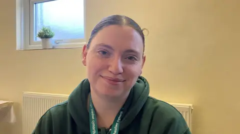 BBC A young woman with brown hair tied back, wearing a green hoodie and sitting at a desk at a Taff Ely Foodbank site. 
