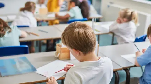 Getty Images Stock image of children with their back to the camera colour do school work. One main child is visible while the rest are out of focus in the background.