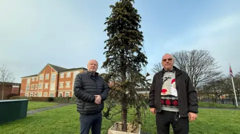 Michael Hartnack and Richard Beck standing next to the vandalised Christmas tree which is about 8ft tall and thin looking. Mr Hartnack is standing to the left of the tree with his hands clasped in front of him. He is wearing a black puffer jacket and has grey hair. Mr Beck is standing on the right of the tree with sunglasses on and a Christmas jumper underneath a dark jacket.