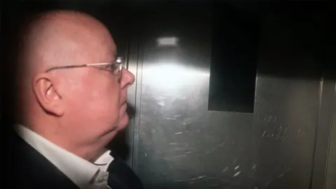 Peter Murrell, who is bald with glasses, photographed in profile in the back of a van. He is wearing a white shirt and black suit jacket, and is illuminated with a flash 