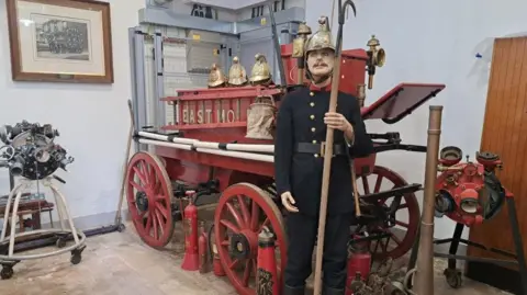 Surrey Fire Museum Firefighting artifacts on display at Surrey Fire Museum, including a wooden fire truck display with a model person standing in front of it in uniform and a gold helmet.