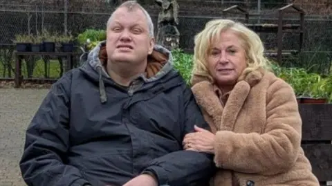 Amanda Shelley Mark Hilton has grey hair and is wearing a blue parka and blue tracksuit bottoms with double white stripe down his legs. Amanda Shelley has long blonde hair, a light brown fur coat, blue jeans and brown cowboy boots.
They are sitting on a bench in a garden nursery.