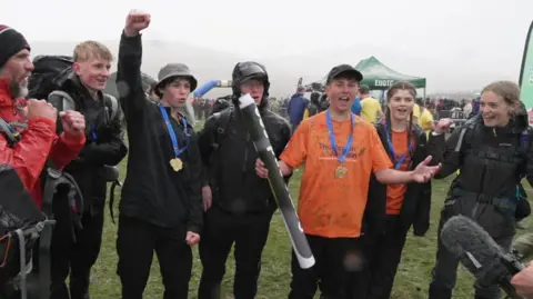 Four young men, two girls and an adult are cheering. The walkers are wearing medals. It is grey and rainy.