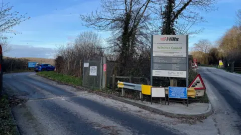 The entrance to Lower Compton household recycling centre near Calne. A sign prominently displays the Hills logo and the Wiltshire Council logo and a blue car can be seen entering the site on the left.