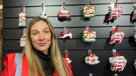 Steve Jones/BBC Sophie Webb, who has long blonde hair and blue eyes. She is standing in front of a variety of Christmas decorations.