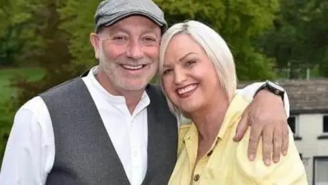 Family photograph Gary wearing a grey flat cap, white shirt and grey waistcoat with his arm around his partner Claire Nutter. She has blond hair and a yellow top. They are both smiling with countryside in the background.
