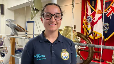 Eliyah is pictured in a navy blue sea cadets polo shirt. She is smiling and has black rimmed glasses. Her hair is dark and tied in a pony tail. She is smiling. 