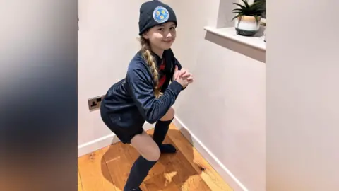 A 10-year-old girl in a football kit demonstrating a squat in a room with white walls and wooden floor. The girl has long fair hair in a plait and is wearing dark socks, shorts and tracksuit top and a red and black striped shirt.