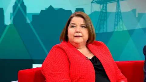 Julie Minns sitting on the Politics North sofa with the teal background behind her.