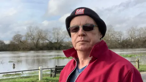 Chertsey resident, Colin Bennett, looks straight at the camera. He is wearing a black hat and a red jacket, and has dark shades on. He is standing in front of a body of water with trees in the background.