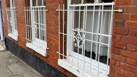 Supplied A property's exterior brick wall with three windows on it. The three windows have thin white metal bars on them. 