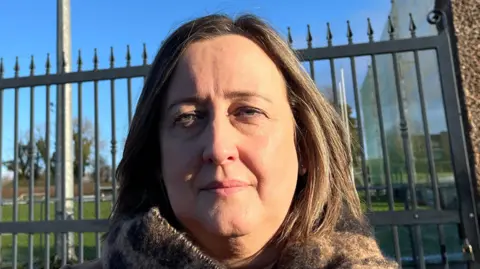 Siobhan Brown standing outside the gates of at Bellaghy GAA ground. She has shoulder-length brown hair and is wearing a brown scarf. The gates of the GAA club can be seen in the background.