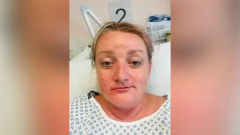 Gabby Rudge-Cox A woman in her forties is sitting in a hospital bed wearing a surgical gown.