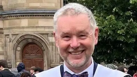 Bill Hepburn looking straight at the camera. He has white hair and a short grey beard. He is smiling. Only his head and shoulders are visible. He is wearing a dark coloured bow tie around the collar of a white shirt.