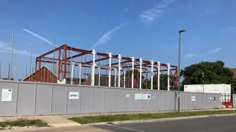 The metal structure of a building under construction behind a grey panel fence