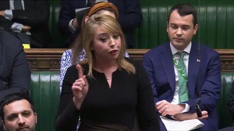 House of Commons Sarah Bool in the house of commons. She is wearing black and is mid-speech. She has one hand in the air with her index finger pointing upwards.