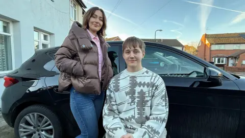 A woman and a teenager in a wheelchair are in front of a black car. They are smiling.