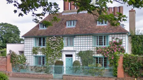 A three-storey village house with a pattern of blue-edged squares on a white background on the front elevation. There is blue door in a white frame. Climbing plants are evident on the facade. There is a small paved area in front of the door with shrubs. There are brick walls either side of the house covered in leaves. A low blue railing separates the property from the road.
