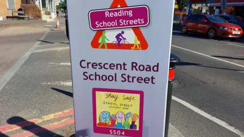 Reading Borough Council A sign attached to a bollard, which reads "Reading school streets" and "Crescent Road school street". On the sign there is also a child's drawing of several smiling people, which reads "stay safe". Behind the sign, there is a black and red bollard. The sign is on a residential road and there are cars in the background.