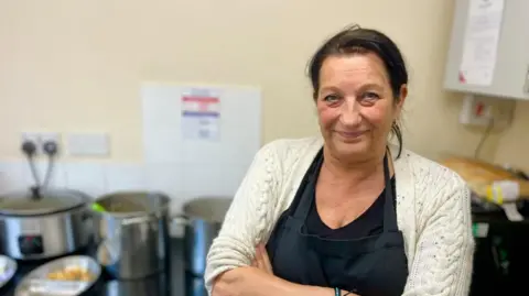 Joanne Beek is standing in a kitchen area wearing a cream-coloured knitted cardigan with a black apron on over a black top. Her arms are crossed. Behind Joanne, there are several large metal cooking pots and a slow cooker on a black counter top.