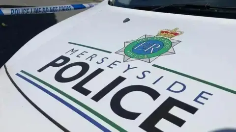 The white car bonnet of a Merseyside Police patrol car, bearing the name of the force in black and blue lettering. Police tape can be seen across a road behind the vehicle.