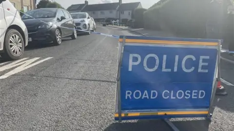 A blue sign which reads 'police road closed' placed on a residential street with a row of cars parked to the left hand side
