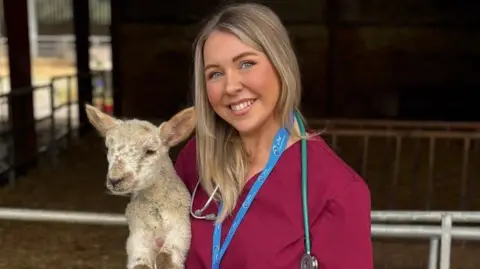 Karis Ramsay holding a lamb in front on an animal pen. Karis is wearing burgundy scrubs, a blue lanyard and ID as well and a stethoscope 