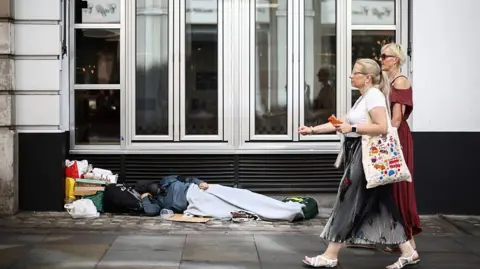 Two women walk past a person lying on the pavement in London, 28 July 