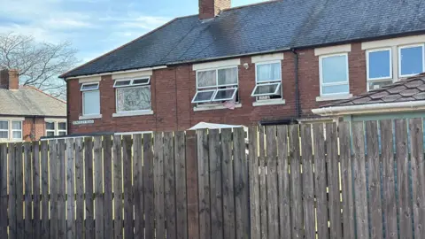 A two-storey terraced house with the upstairs windows wide open. Pink curtains can be seen hanging out one of the windows.