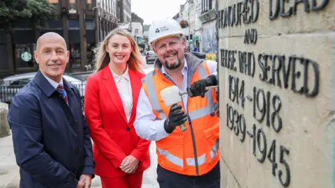 DCSDC Two men and a woman are standing beside the war memorial in Derry.
Anne Carlin is wearing a red suit with white shirt. Johnny Booth is wearing a high-vis vest and Jason Dougherty is wearing a navy overcoat and striped tie with white shirt. 