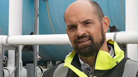 Mark Manton looks toward the camera. He is wearing a yellow high-vis jacket and is standing in front of pipes at Jersey's desalination plant.