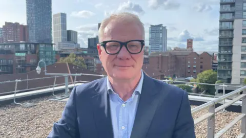 Richard Parker. A man with short grey hair, black glasses, a light blue shirt and navy blazer is standing on a balcony with the Birmingham city centre skyline behind him.