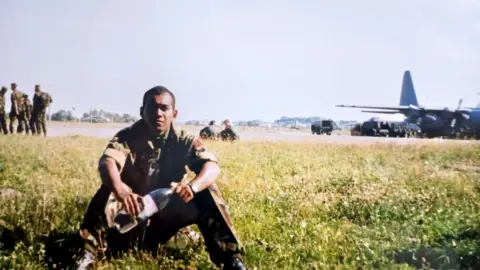Royal British Legion A soldier sitting on a field. Behind him are more soldiers and a warplane.