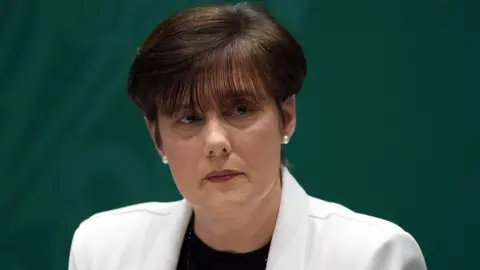 Niall Carson/PA Wire Norma Foley is wearing a white jacket and pearl earrings. She has a black top on. Her hair is brown and bobbed. A backdrop behind her is dark green. 