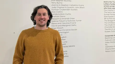 Greg Norrington, programmer at the De La Warr Pavilion, is standing in front of names listed on a white wall in the venue. He is wearing a jumper and T-shirt and smiling at the camera.
