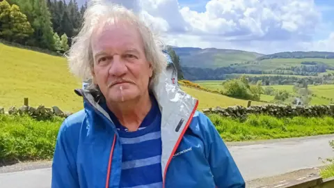 Derek Angelis A man with longish grey hair and a small beard in a blue rain jacket with a light blue and darker blue striped jumper underneath. He is standing next to a pathway with the rolling Borders hills behind him.